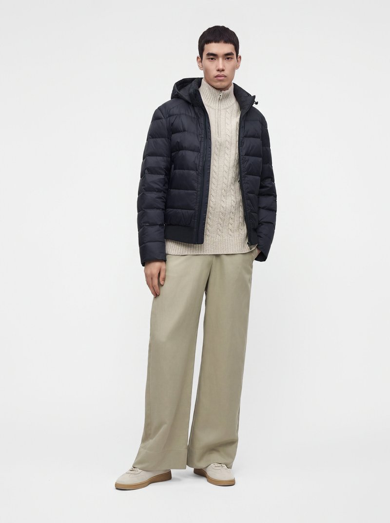 Jeune homme portant une doudoune noire sur un pull blanc en maille torsadée, un pantalon beige à jambes larges et des baskets blanches, debout devant un fond blanc.