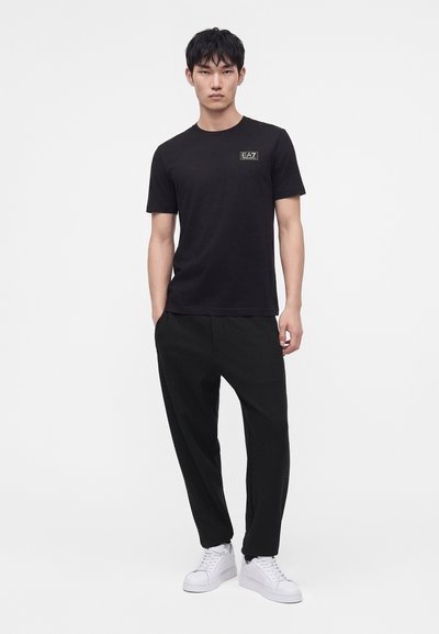 Νέος άνδρας που στέκεται, φοράει μαύρο t-shirt EA7, φαρδιά μαύρη παντελόνια και λευκά αθλητικά παπούτσια σε απλό φωτεινό φόντο.