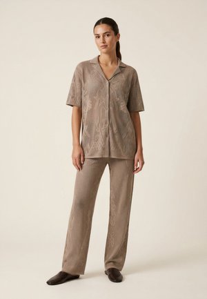 Femme portant une chemise boutonnée beige texturée à manches courtes et un pantalon large assorti, debout devant un fond uni.