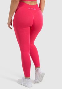 Růžové legíny s vysokým pasem mají elegantní, pružnou tkaninu s hladkým povrchem, doplněné malým logem na pasu. Bílí tenisky dotvářejí celkový vzhled.