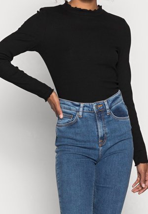 Femme portant un haut noir à manches longues avec des bords volantés et un jean bleu taille haute, main reposant sur la hanche sur fond clair.