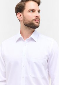 Camicia bianca con colletto a punta, taschino frontale e tessuto liscio con una leggera lucentezza. Una cucitura dettagliata è visibile lungo le cuciture.