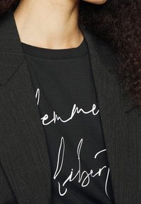 Camiseta negra con texto en cursiva blanca, llevada debajo de una chaqueta gris oscura de tela texturizada. Primer plano del cuello y el hombro.