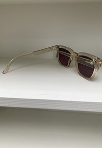 Transparante rechthoekige zonnebril met donkerpaarse lenzen, metalen accenten en gebogen veren. Gegraveerd merk op de pootjes.