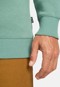 Felpa verde menta con polsini e orlo a coste, caratterizzata da un'etichetta in tessuto sul lato. La mano è appoggiata su una superficie marrone texture.