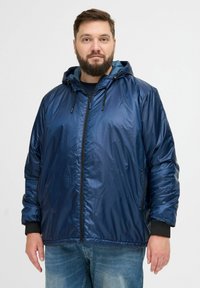 Temno modra jakna s kapuco iz sijočega, lahkega materiala. Ima sprednji zadrgo, nastavljivo kapuco in elastične manšete.