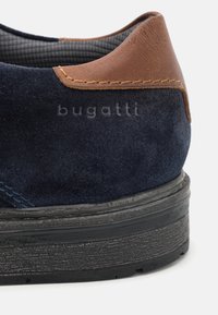 Talons de chaussures en daim bleu foncé avec garniture en cuir marron et semelle noire texturée, avec marquage en relief "bugatti" sur le côté.