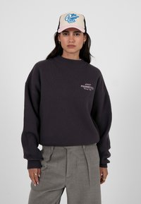 Sweatshirt gris foncé surdimensionné avec col rond et logo brodé "PROHIBITED". Associé à un pantalon plissé gris et une casquette crème ornée de motifs bleus.
