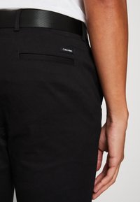 Pantaloni neri realizzati in un tessuto liscio, con una tasca posteriore decorata con un'etichetta Calvin Klein e una cintura nera elegante. La vestibilità è su misura e affusolata.