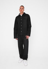 Zwart denimjack met knoopsluitingen, uitgerust met een kraag en een borstzak, gecombineerd met bijpassende zwarte denimbroek en witte sneakers.