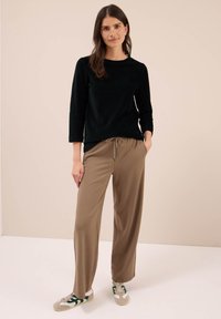 Vrouw met lange bruine haren, gekleed in een zwart lange mouw top, losse bruine broek en witte sneakers met groene accenten, staande in een neutrale ruimte.