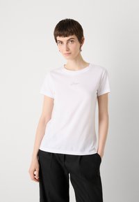 White short-sleeve t-shirt in soft cotton with a round neckline and a subtle logo detail on the chest. Paired with black trousers.