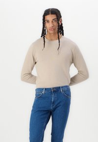 Pulover tricotat bej cu guler rotund, purtat cu blugi denim albaștri. Tesătura este netedă, iar croiala este lejeră și casual.