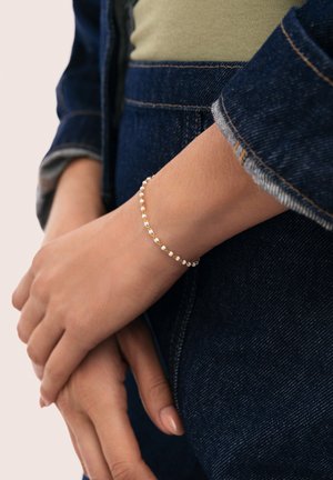 Delicada pulsera de oro con pequeñas cuentas blancas llevada en la muñeca izquierda sobre chaqueta y pantalones vaqueros, manos suavemente entrelazadas.