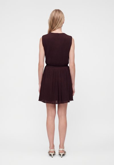 Femme aux cheveux blonds portant une robe plissée violette foncée sans manches et des sandales à talons beiges, debout de dos sur un fond uni.