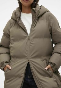 Veste matelassée verte olive, dotée d'une fermeture éclair sur le devant, d'une capuche et de poignets élastiques. Forme structurée avec des poches latérales et une texture de tissu lisse.