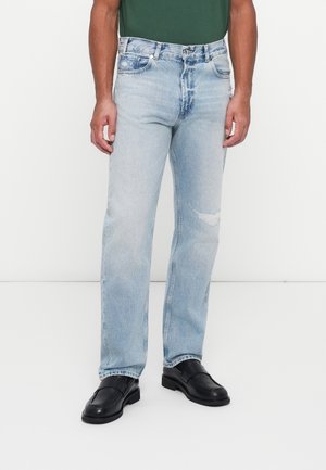 Homme portant un jean droit bleu clair avec une petite déchirure à un genou, associé à des mocassins en cuir noir et un haut vert foncé.