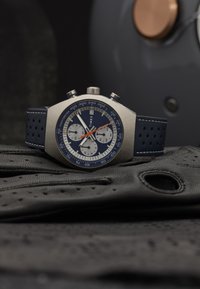 Timex chronograafhorloge met een roestvrijstalen kast, donkerblauwe wijzerplaat, witte subdials, oranje accenten en een marineblauwe leren band op zwarte handschoenen.