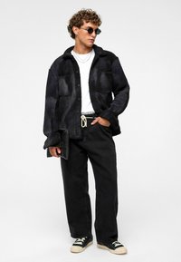 Fluffy zwarte colbert met twee borstzakken, gedragen over een witte blouse. Gecombineerd met losse zwarte broek en zwarte sneakers met crème accenten.