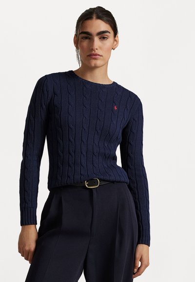 Granatowy sweter w warkoczowym splocie z okrągłym dekoltem i długimi rękawami, z małym czerwonym logo. Połączony z ciemnymi spodniami o wysokim stanie.