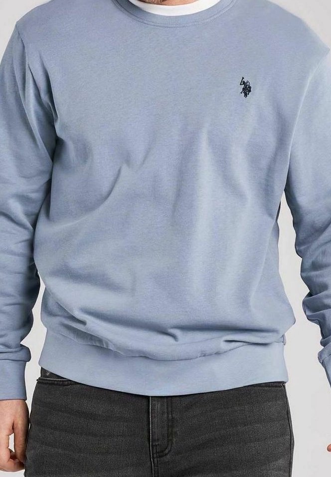 Homme portant un sweat-shirt bleu clair à manches longues avec un petit logo noir de joueur de polo sur la poitrine et un jean gris foncé.