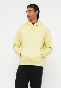 Mlad oseba z kite, oblečena v svetlo rumeno kapuco Adidas, z rokama v sprednjem žepu, gleda na stran.