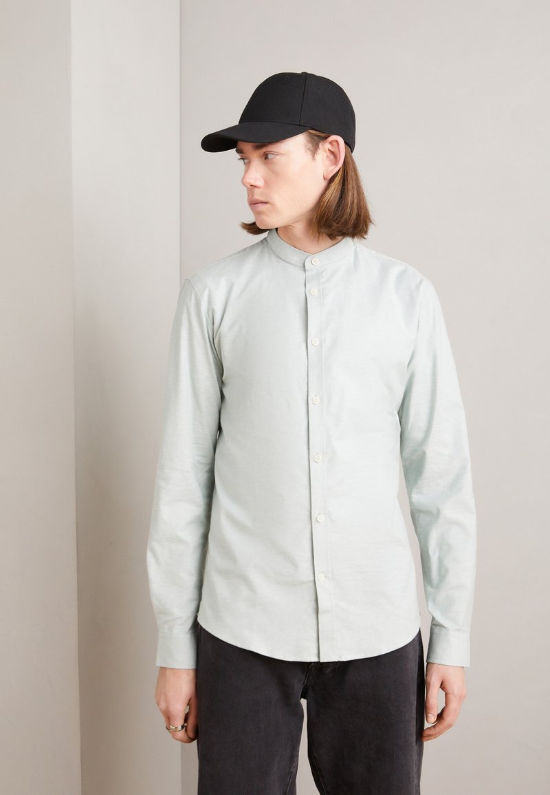 Camicia button-up azzurro chiaro con colletto alto, maniche lunghe e una texture liscia. Abbinata a un cappellino nero a tinta unita e pantaloni scuri.
