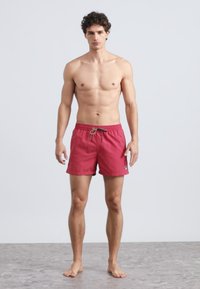 Röda badshorts av lätt tyg med elastisk midja och dragsko samt en liten logga på fållen.