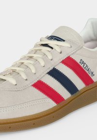 Μπεζ σουέντ αθλητικό παπούτσι Adidas Spezial με κόκκινες και ναυτικές ρίγες, λευκά κορδόνια και σόλα από καουτσούκ.