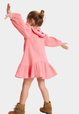 Jong meisje in een roze jurk met kersenborduursel op de kraag, draagt bruine laarzen met franjes, armen uitgestrekt, gezicht naar achteren gericht.