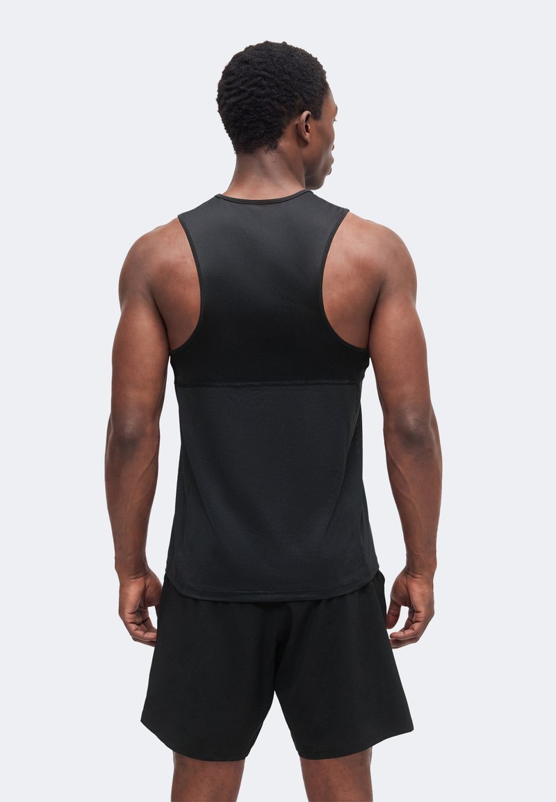 Homme musclé debout de dos, portant un débardeur de sport noir et un short noir sur un fond uni clair.