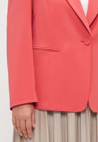 Korallfarget blazer for kvinner med knappelukking, en falsk lomme og glatt stoff. Båret over en plisserte, beige, blank nederdel.