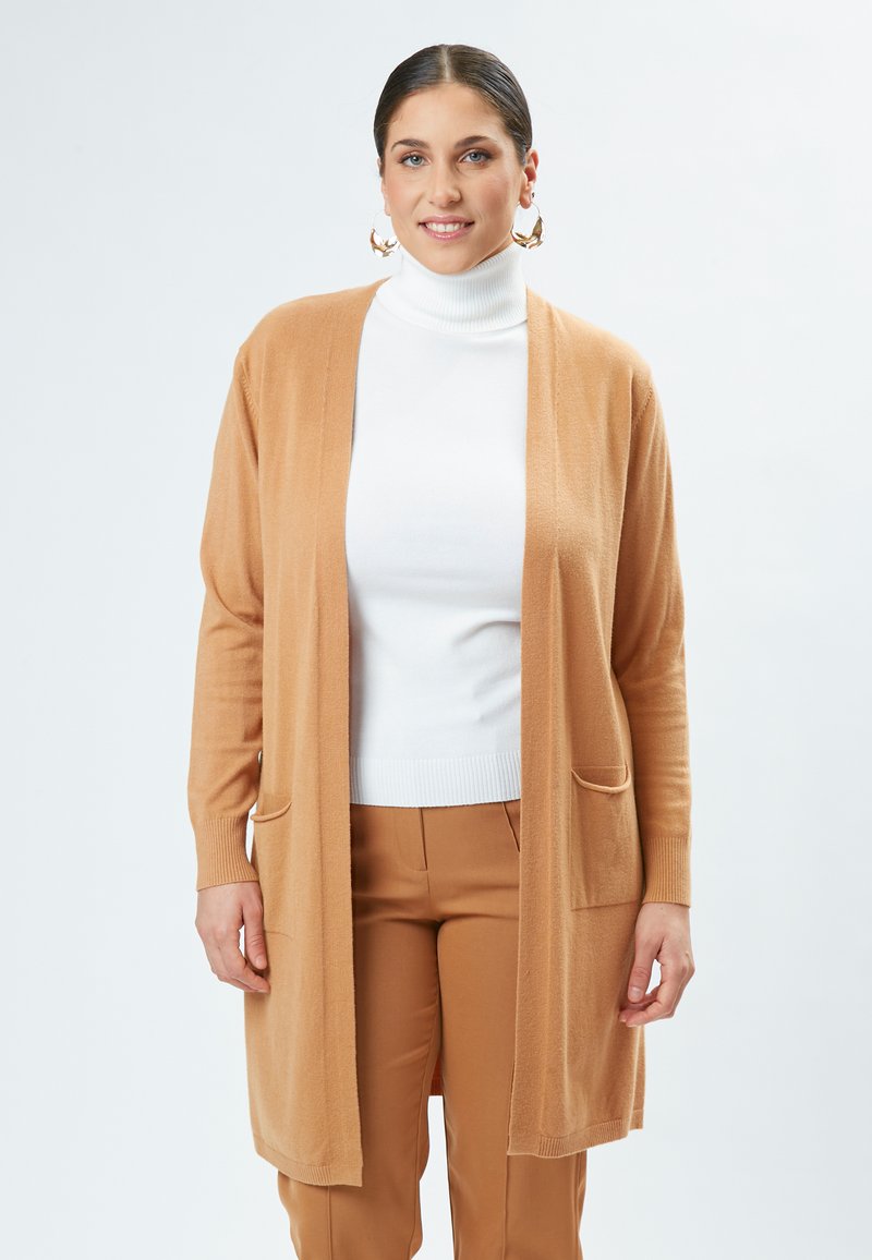 Cardigan lungo a frontale aperto di colore marrone chiaro, realizzato in tessuto morbido con due tasche frontali. Indossato sopra un dolcevita bianco.