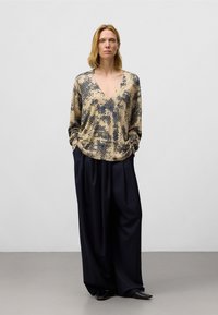 Beige blouse met een patroon van grote marineblauwe stippen, met een V-hals en lange mouwen. Gecombineerd met losse, marineblauwe broek met brede pijpen en zwarte schoenen.