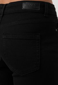 Black denim jeans featuring a smooth texture, stitched detailing, and a leather brand patch near the waistband. Back pocket visible.