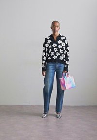 Musthave mustreid valge lillemuster, sinised teksad, metallikhõbedased kingad, hoides mitmevärvilist kotti, millel on tekst "KOTT".