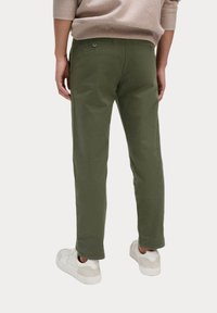 Olivengrüne Baumwoll-Chinos mit schlanker Passform, zwei Gesäßtaschen und Nahtdetails, kombiniert mit weißen Sneakern und einem hellbeigen Sweater.