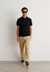 Črna polo majica s kratkimi rokavi, khaki hlače in temna sončna očala. Model hodi proti enotni ozadju, slikovito predstavlja priložnostno obleko.