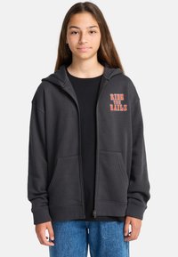Jongere met lang bruin haar, gekleed in een zwarte hoodie met ritssluiting waarop de tekst "RIDE THE RAILS" staat, en blauwe spijkerbroeken, staand tegen een effen achtergrond.