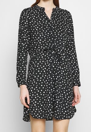 Sort langærmet kjole med hvide uregelmæssige polkaprikker, knapper foran, bindebånd i taljen og en let buet kant over knæene.