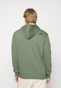 Felpa verde oliva realizzata in morbido tessuto, con cappuccio regolabile, polsini a costine e un logo sulla spalla sinistra. Design semplice.