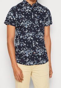 Camicia a maniche corte con bottone di colore navy, decorata con un motivo floreale bianco e azzurro; presenta un colletto e una tasca, realizzata in tessuto leggero.