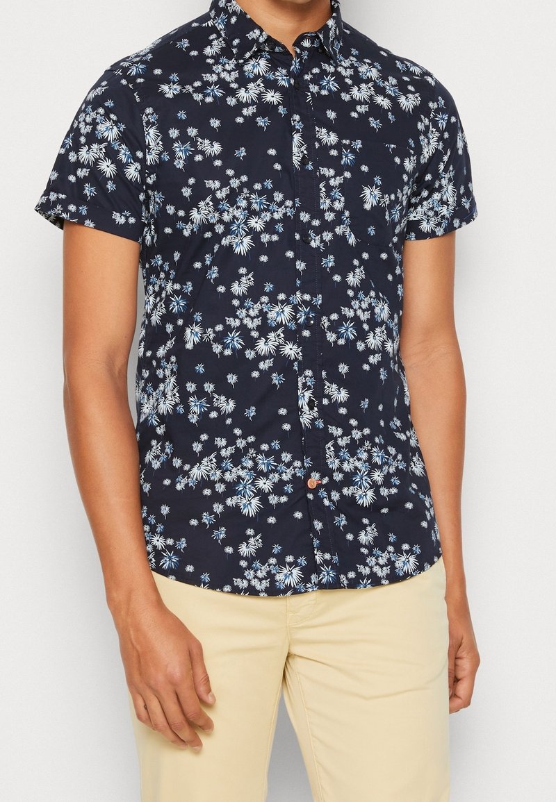 Camicia a maniche corte con bottone di colore navy, decorata con un motivo floreale bianco e azzurro; presenta un colletto e una tasca, realizzata in tessuto leggero.