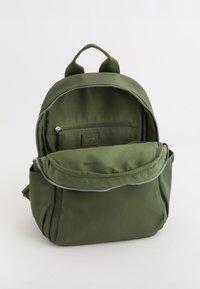 Mochila de color verde oliva hecha de tela duradera, con un compartimento principal con cremallera, bolsillo frontal y asas de tela dobles. Diseño compacto.