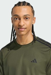 Olivovozelené športové tričko s textúrovaným vzorom, čierne trojprúžkové akcenty na pleciach a malé čierne logo na hrudi.