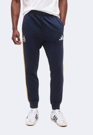 Bărbat purtând pantaloni sportivi bleumarin cu dungi galbene, cu logo-urile Real Madrid și Adidas, asortați cu pantofi sport albi Adidas cu dungi negre.