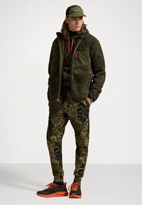 Olivgrüne Fleecejacke mit orangen Akzenten, schwarzer Pullover mit Muster, passende Hose und schwarze Schuhe mit orangen Details. Kappe inbegriffen.