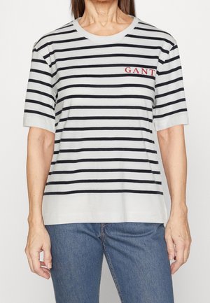 Person iført en hvid og sort vandret stribet kortærmet skjorte med rød "GANT" logo på brystet og blå jeans.