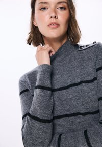 Maglione grigio con strisce orizzontali nere, colletto alto e dettagli con bottoni sulla spalla. Materiale a maglia morbido con design aderente.