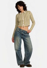 Γυναίκα που φοράει ένα κοντό, ριγέ πουκάμισο με κουμπιά και φαρδιά τζιν με ξεθωριασμένο σχέδιο, με καφέ παπούτσια, στέκεται με το ένα χέρι στην τσέπη μπροστά σε λευκό φόντο.